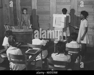 A photograph of two girls giving a presentation at the front of their class room, their display is titled 'A Health Moving Picture', the girl on the right uses a pointing stick to direct the attention of her classmates while her partner operates the display, the teacher watches their presentation from behind her desk on the left side of the classroom, the other students in the room are also girls in the 4th and 5th grade of the Flint River School in Georgia, 1939. From the New York Public Library. Stock Photo