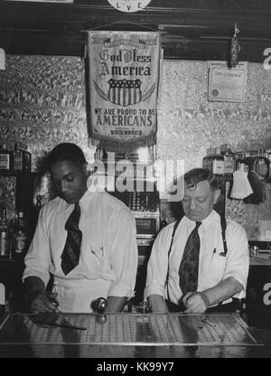 A photograph of two men working behind a bar, the bartenders are pouring beers from a set of taps that has been installed below the level of the bar, a stainless steel drip tray stands in front of the them, both men wear button down white shirts and ties, a large banner that reads in part 'God Bless America' and their City of Chicago business licenses hang on the wall behind them, Southside of Chicago, Illinois, 1940. From the New York Public Library. Stock Photo