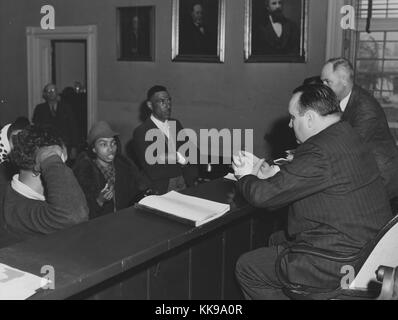 A photograph of a group of African American citizens appearing before two white court officials, everyone is dressed in nice clothing, the two groups are separated by a high wooden barrier and the officials are on an elevated platform, another man watches from a doorway, the room is decorated with three portraits of different men, Rustburg, Virginia, March, 1941. From the New York Public Library. Stock Photo