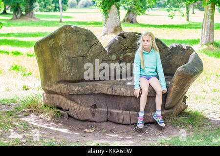 Child sitting on a large bench / seat carved from wood looking pensive / thoughtful on a bright sunny day, Ireland little girl sitting bench Stock Photo