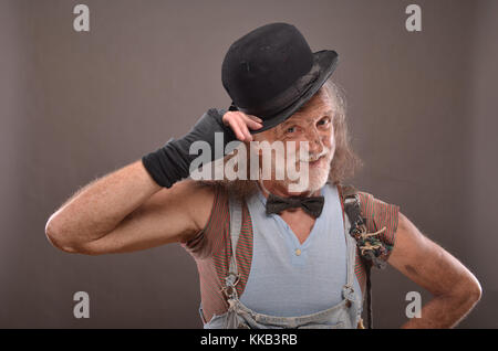 Happy old beggar, smiling and touching his black bowler hat, posing against gray background Stock Photo