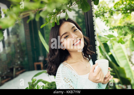 Happy young asian woman with mug in hands drinking coffee standing on balcony. Stock Photo
