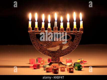 Hanukkah menorah with burning candles sits on the table; the lights reflect in the dark window behind them; dreidels scattered on the table in front o Stock Photo