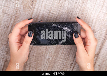 A smartphone with a broken screen in the girl's hand on a wooden table. Close-up. Studio lighting. Stock Photo