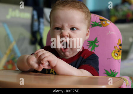 Portrait of yawning redheaded baby with big blue eyes and opened mouth dressed in blue shirt, in the room. Stock Photo