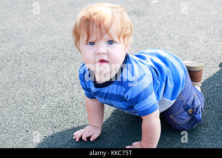 Fashioned ginger toddler boy with blue eyes staying on all fours on the play ground and watching on camera. Stock Photo