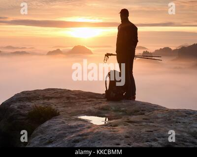 Tired hiker with sporty backpack on rocky peak  and watching into deep misty valley bellow. Sunny spring daybreak in rocky mountains. Stock Photo