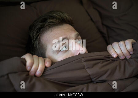 Caucasian young man hiding in bed under the blanket at home. Stock Photo