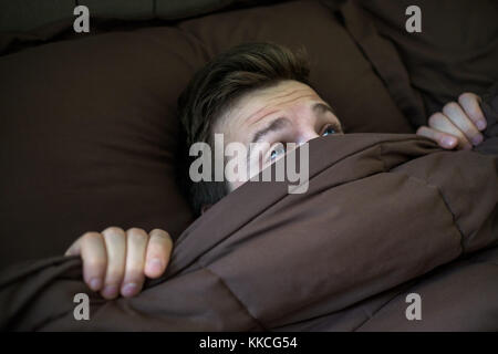 Caucasian young man hiding in bed under the blanket at home. Stock Photo