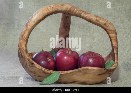 Three red apples in wooden basket with few green leaves on rough natural sacking material. Unfocused background. Stock Photo