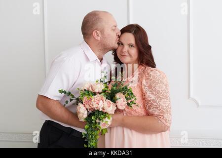 Man kissing smiling  pregnant woman in coral dress  who holding bouquet of rose peonies. Happy kissing couple  on luxury decorated white wall. Stock Photo