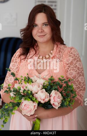 Beautiful pregnant woman with happy smiling face and brown hair dressed in coral dress and jewelry holding bouquet of rose peonies in luxury decorated Stock Photo