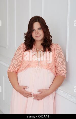 Beautiful pregnant woman with calm smiling  peaceful face dressed in coral dress and jewelry embracing her belly standing on white luxury decorated wa Stock Photo