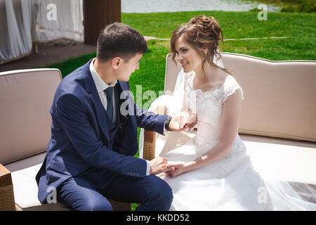 Groom sitting with young bride on sofa in nature and holding her hand Stock Photo