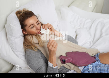 Sick woman with a miserable expression taking her temperature as she lies in bed suffering from a seasonal cold and flu trying to keep warm
