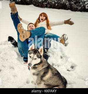 Cheerful couple is having fun by laying on the snow while the siberian husky is sitting in the front of them. Funny portrait. Stock Photo