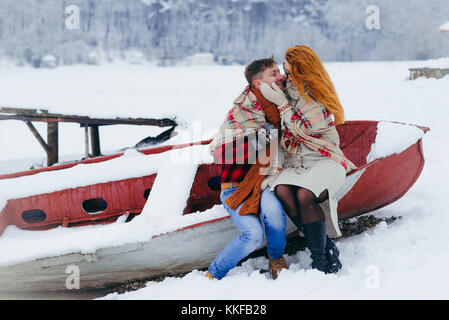 Horizontal winter portrait of the cheerful loving couple. The charming woman is softly stroking the cheek of the man while sitting on the boat. They are wrapped in the plaid during the snowfall. Stock Photo