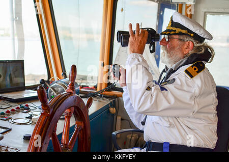 Old experienced captain observes using binoculars by left hand and holding radio comunication equipment by right hand Stock Photo