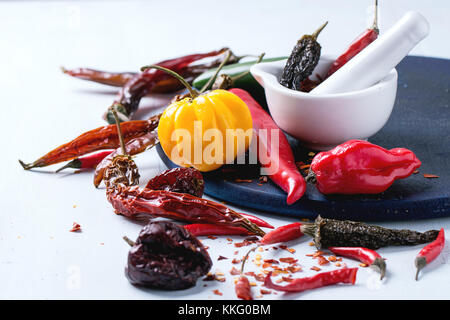 Assortment of fresh, dryed and flakes hot chili peppers with white ceramic mortar on dark blue cutting board over light blue wooden background Stock Photo