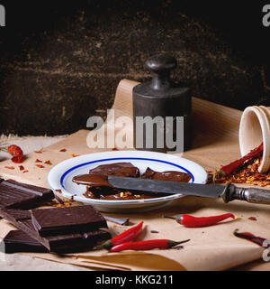 Chopping dark chocolate with fresh and dry red hot chili peppers on baking paper with plate of hot melted chocolate and vintage knife. Stock Photo