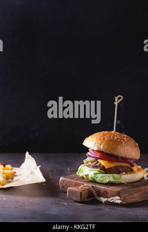 Fresh homemade burger on little wooden cutting board and grilled potatoes over dark background. With copyspace on top Stock Photo