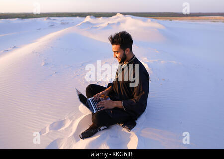 Handsome young Arab man writes on Internet using laptop and sits Stock Photo