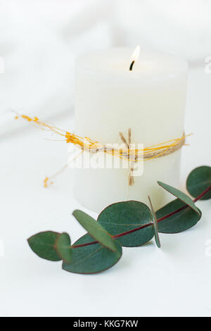 Thick White Lit Candle with Twine and Dry Plant Twig Silver Dollar Eucalyptus White Cloth Background Copy Space. Banner Poster Postcard Template Inspi Stock Photo