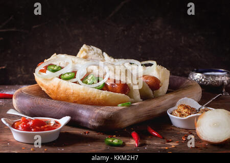 Homemade hot dogs on wooden plate with ingredients mustard, tomato sauce, onion, pepper, rosemary on wooden table. Dark rustic style Stock Photo