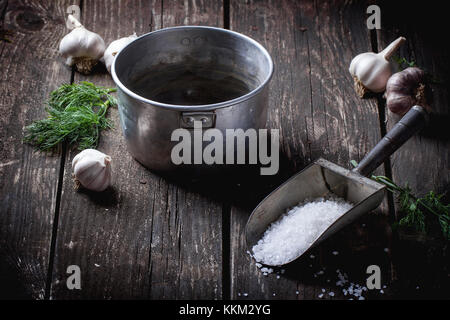 Old aluminum pan for preparation low-salt pickled cucumbers. Over black wooden table with sprinkled sea salt, dill and garlic. Dark rustic style. Stock Photo