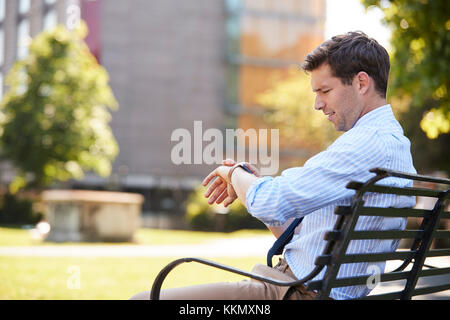 Businessman Sitting In City Park Looking At Smart Watch Stock Photo