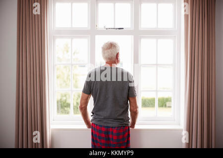 Rear View Of Senior Man In Pajamas Looking Out Of Bedroom Window Stock Photo