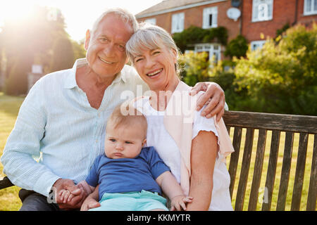 Grandparents Sitting On Seat In Garden With Baby Grandson Stock Photo