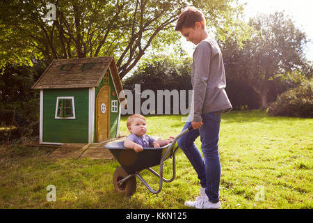 Boy Pushing Baby Brother In Garden Wheelbarrow Stock Photo
