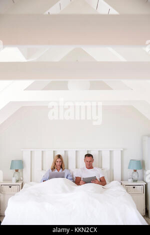 Couple Wearing Pajamas Lying In Bed Using Digital Devices Stock Photo