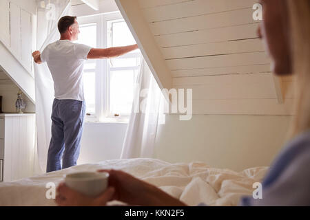 Man Opens Curtains And Looks Out Of Window As Woman Lies In Bed Stock Photo