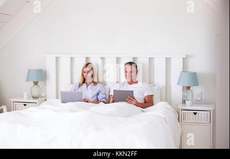 Couple Wearing Pajamas Lying In Bed Using Digital Devices Stock Photo
