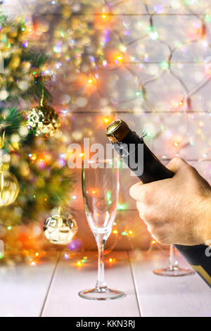 Men's hands open a bottle of champagne on the background of a New Year tree and garlands and glasses Stock Photo