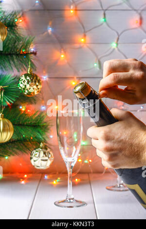 Men's hands open a bottle of champagne on the background of a New Year tree and garlands and glasses Stock Photo