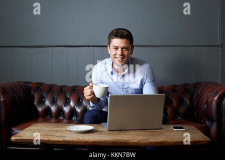 Businessman Drinking Coffee Whilst Working On Laptop In Internet Cafe Stock Photo