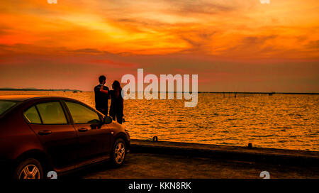 Silhouette of happiness couple standing by the car at the seaside at sunset. Beautiful orange sky and clouds Stock Photo