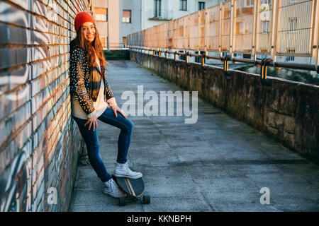 Young  and beautiful skater woman enjoying the sunset in the city resting on a brick wall with graffiti Stock Photo