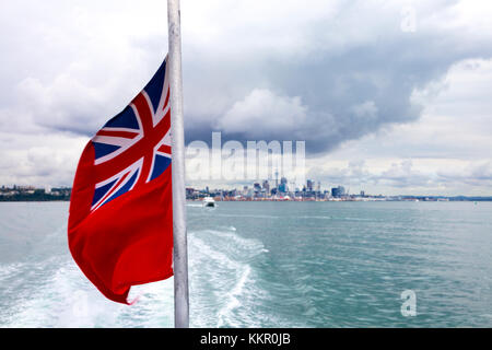 Red ensign flag at the stern of a boat waving in the wind with skyline of Auckland in the background, New Zealand Stock Photo