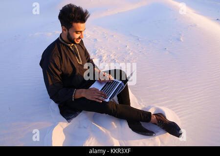 Young and handsome Muslim guy sits in social networks using computer, prints message or reads news, rests from turmoil of city, enjoys tranquility of  Stock Photo