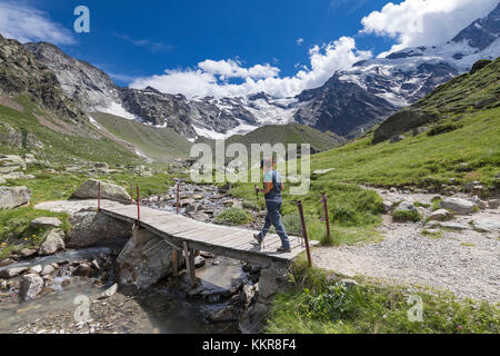 A trekker crosses a small wooden bridge near the Zamboni Zappa refuge at the foot of the East face of Monte Rosa Massif (Macugnaga, Anzasca Valley, Verbano Cusio Ossola province, Piedmont, Italy, Europe) Stock Photo