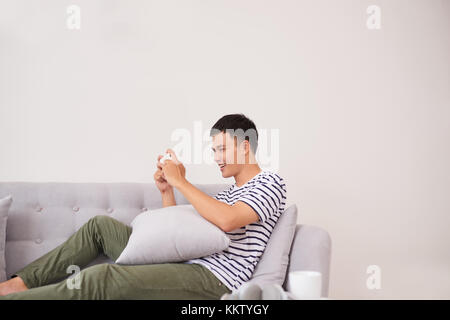 Young asian man sitting on sofa and playing games on phone at home Stock Photo