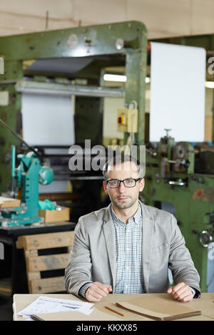 Young factory worker in casualwear sitting by table in industrial workshop Stock Photo