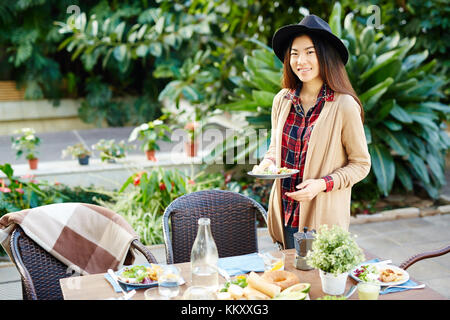 Happy girl with long hair holding plate with fresh food while standing by served table Stock Photo