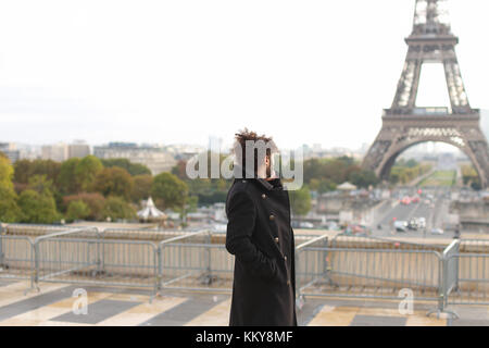Half Nigerian guy near Eiffel Tower sends messages to girlfriend using modern smartphone in slow motion. Handsome jocund man wears black coat, ring wi Stock Photo