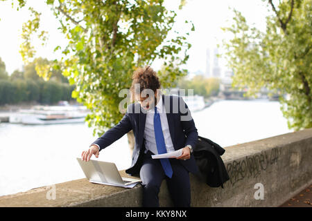 Foreign academician visiting France for scientific conference and working near river with laptop. Young man dressed in black coat working with laptop  Stock Photo