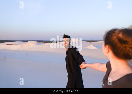 Handsome male Arab holding woman's hand, chatting and smiling looking at her. Lovers walk through expanses of sandy desert at sunset under open blue s Stock Photo